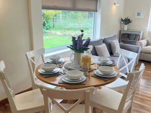 A dining area with a round table set for four, surrounded by white chairs, with a view of greenery outside. - Saddleback Bungalow (Llandrindod Wells)