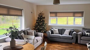Ein Wohnzimmer mit zwei grauen Sofas, einem Holztisch, einem weißen Sessel, einem Weihnachtsbaum und einem Fenster mit Jalousien.