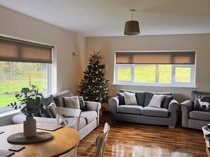 Ein Wohnzimmer mit zwei grauen Sofas, einem Holztisch, einem weißen Sessel, einem Weihnachtsbaum und einem Fenster mit Jalousien.