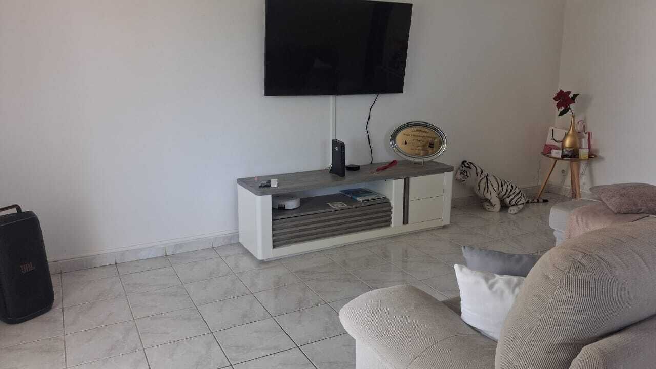 Un salón con televisor de pantalla plana montado en la pared, un mueble blanco para equipo de sonido con un reloj, un sofá cómodo y una mesita con adornos.
