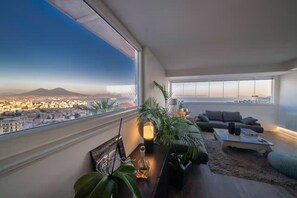 Ein modernes Wohnzimmer mit einem großen Fenster, das einen Blick auf eine Stadtlandschaft und ein Gebirge bietet.