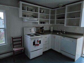 A compact kitchen with white cabinets, a stove, a sink, and a chair.