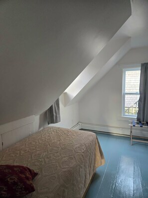 Ein Schlafzimmer mit schrägem Dach, einem Bett mit gemustertem Überwurf, einem Fenster mit Vorhängen und einer Heizkörper.