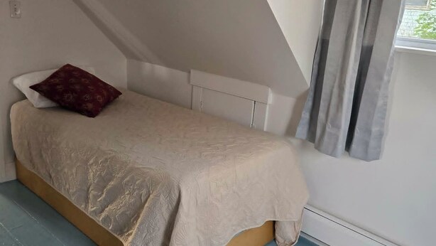 Une petite chambre avec un lit simple, un plafond en pente, une fenêtre avec des rideaux et un radiateur.