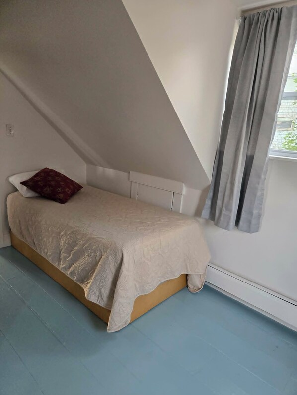 A small, single-bed room with a sloped ceiling, a window with curtains, and a radiator.