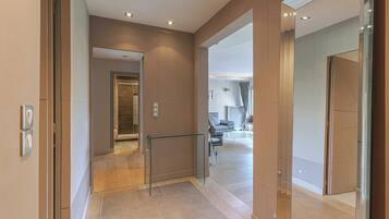 A modern interior with a hallway leading to a living area, featuring a glass partition, a staircase, and a visible elevator.