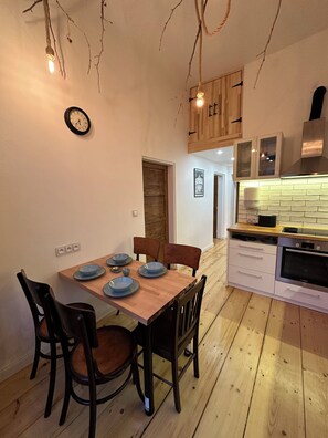 Un comedor con una mesa de madera dispuesta para cuatro personas, rodeada de sillas. Se ve una cocina con electrodomésticos y armarios modernos al fondo.