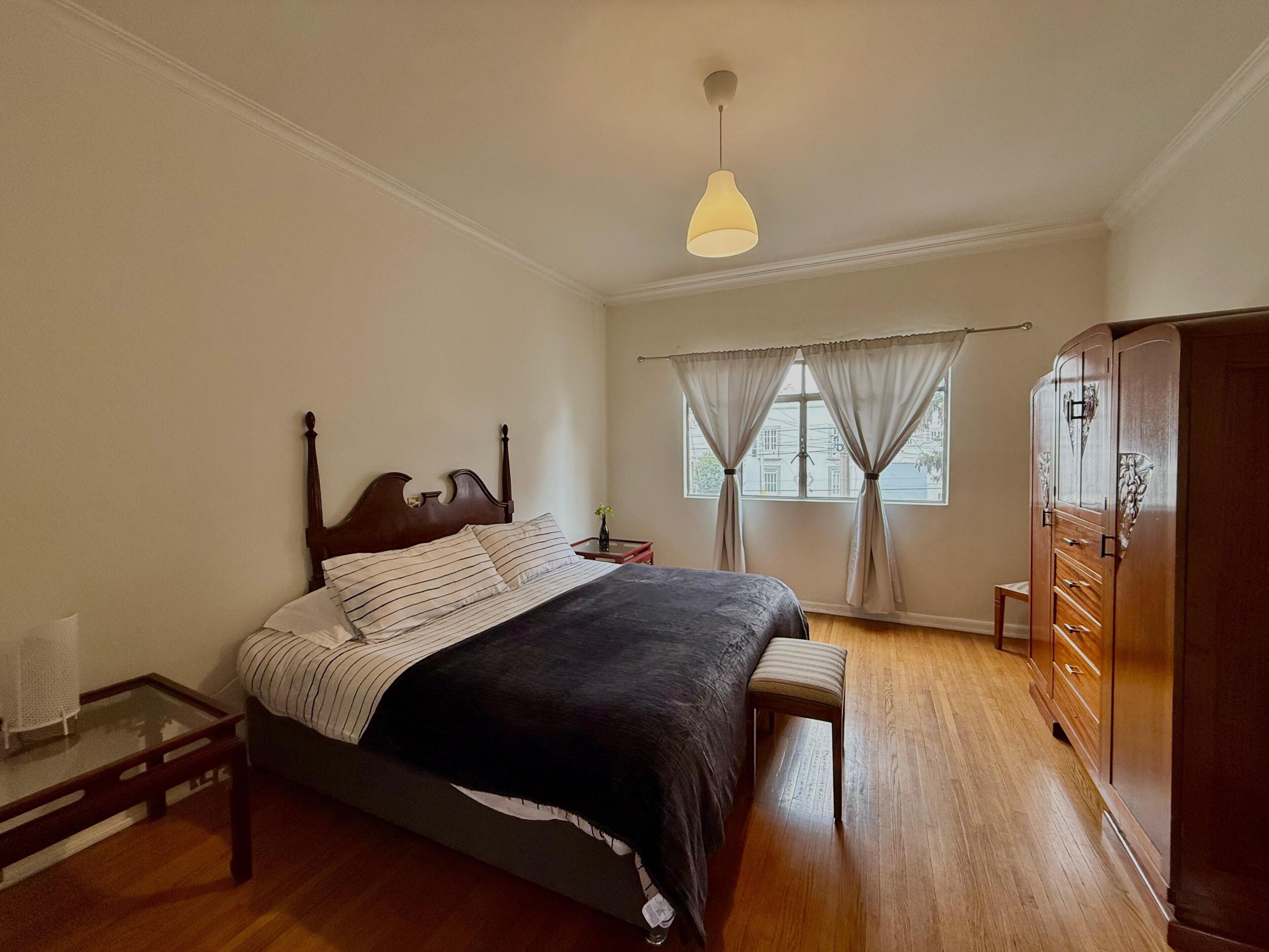 A bedroom with a wooden bed, a bedside table, a nightstand, a wardrobe, and a window with curtains.