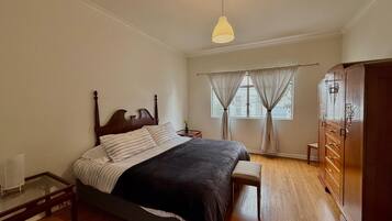 A bedroom with a wooden bed, a bedside table, a nightstand, a wardrobe, and a window with curtains.