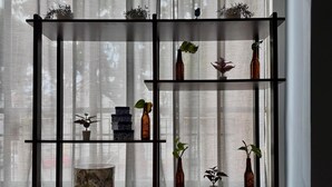 Una habitación con piso de madera, una estantería metálica negra con diversos objetos y cortinas de tela ligera.