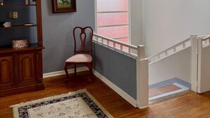 Una habitación con una silla de madera, una estantería, un cuadro enmarcado, un ventanal con vidrio esmerilado y una araña.