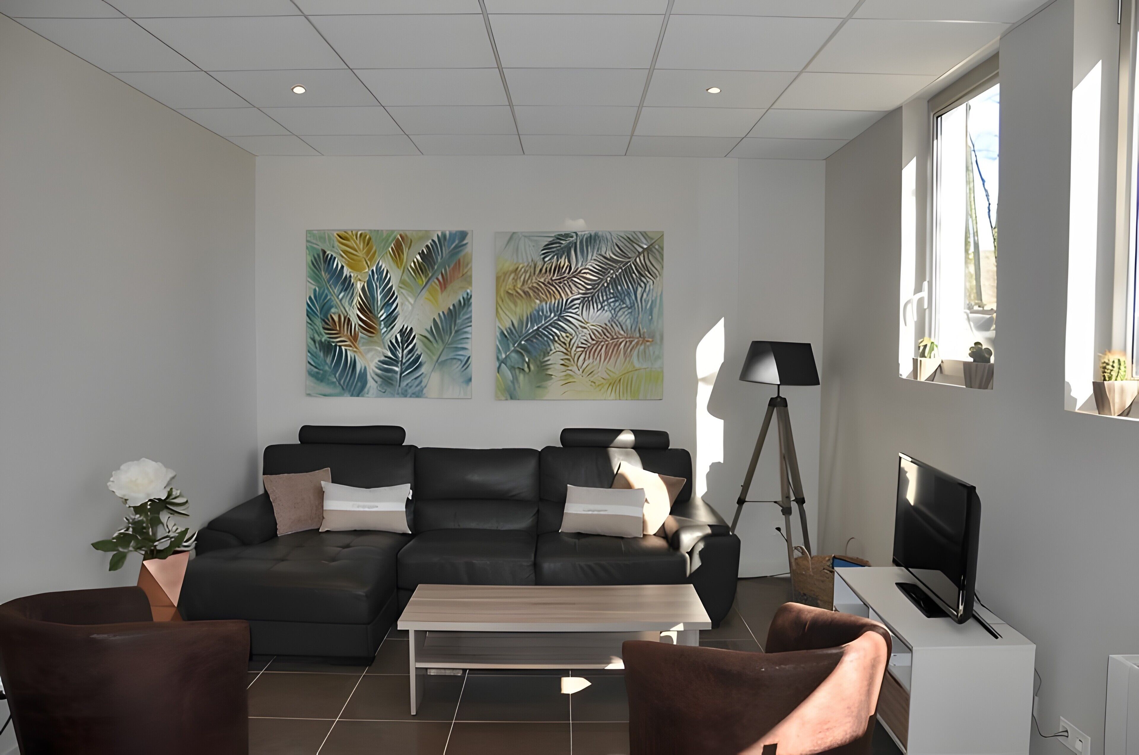 Een moderne woonkamer met een hoekbank, een salontafel en een televisie. Twee grote schilderijen hangen aan de muur, er staat een potplant en een vloerlamp.
