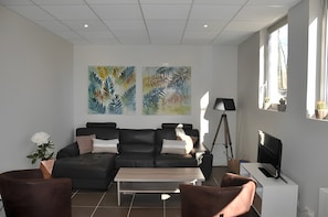 A modern living room with a sectional sofa, a coffee table, and a TV. There are two large paintings on the wall, a potted plant, and a floor lamp.