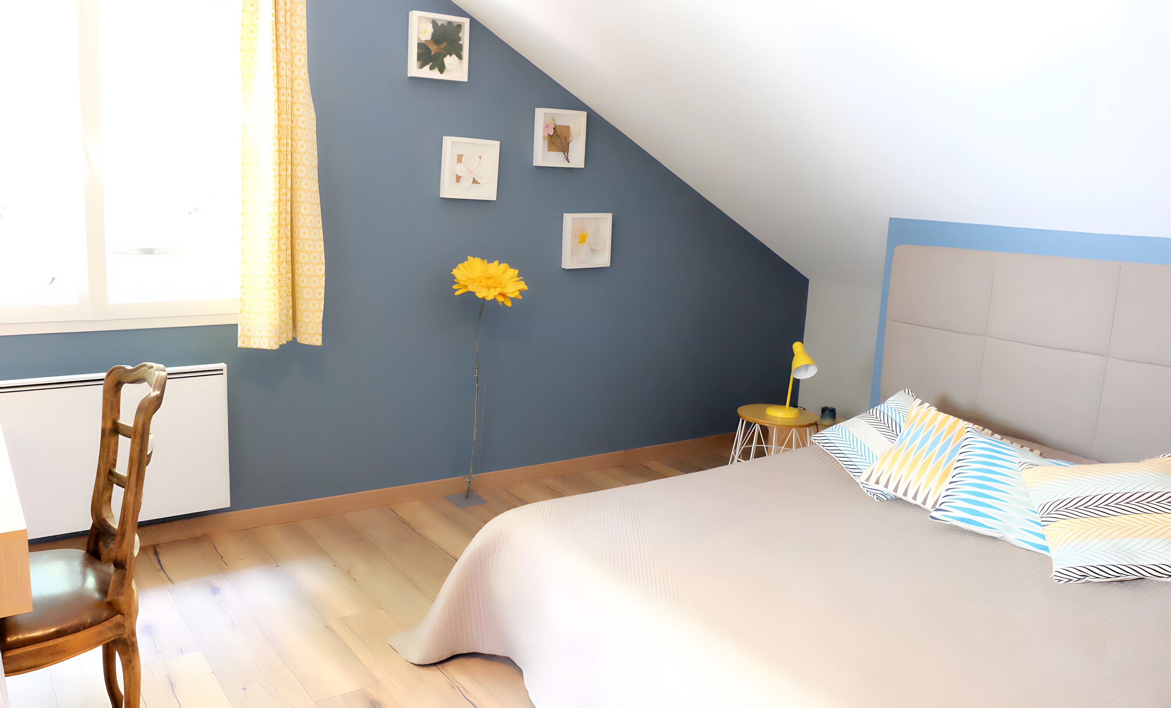 Une chambre avec un plafond en pente, un lit avec des coussins à rayures, un bureau, une chaise et une fenêtre avec des rideaux.