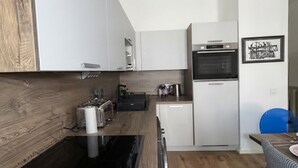 Moderni keittiö, jossa on valkoiset kaapit, sisäänrakennettu uuni, kahvinkeitin ja ruokailutila, jossa on sininen tuoli.