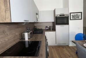 A modern kitchen with white cabinetry, a built-in oven, a coffee machine, and a dining area with a blue chair. - celine 36 (Paris)