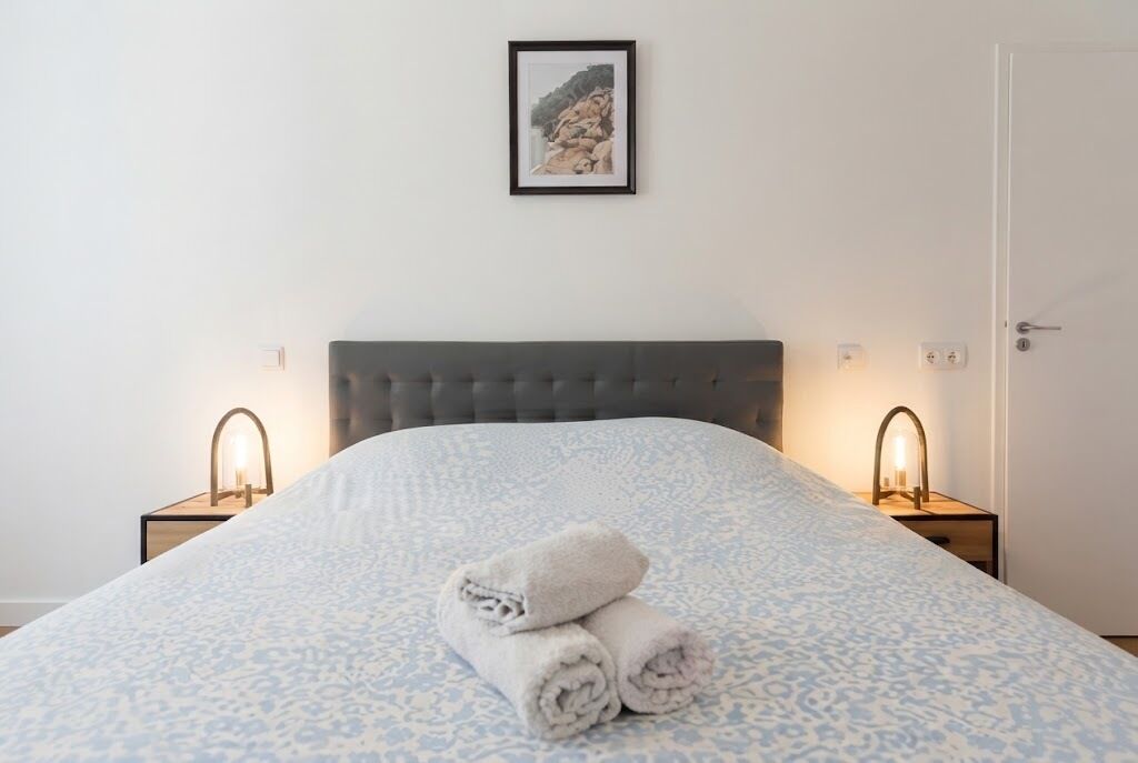 Uma cama bem arrumada com colcha estampada e toalhas dobradas, duas luminárias de cabeceira e um quadro na parede.