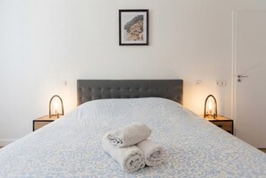 A neatly made bed with a patterned bedspread and rolled towels, two bedside lamps, and a framed picture on the wall. - celine 36 (Paris)