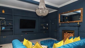 A living room with a large blue sofa, a flat-screen TV, a chandelier, and a fireplace. - Kexby House (York)