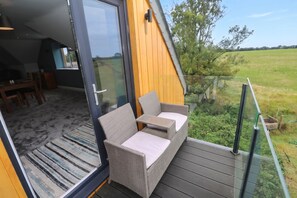 Un balcón con un sofá y una mesa de mimbre, un ventanal de vidrio que da a un espacio interior y vistas a un campo verde.