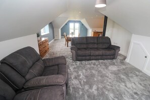 Ein Dachzimmer mit grauem Teppich, zwei dunklen Sofas, einer Holzkommode und einem Esstisch mit Stühlen.