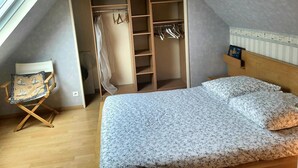 A bedroom with a sloped ceiling, a bed with floral bedding, a wooden wardrobe with hanging space, and a chair with a cushion. - Villa 'Front De Mer Les Cordylines' with Sea View, Private Terrace and Wi-Fi (Saint-Germain-sur-Ay)