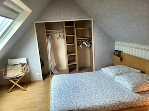 Un dormitorio con techo inclinado, una cama con ropa de cama floral, un armario de madera con espacio para colgar y una silla con cojín.
