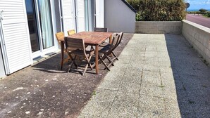 A patio with a table and chairs, a building with windows, and a hedge in the background.
