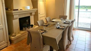 A dining area with a fireplace, a table set for six, and a view through a glass door. - Villa 'Front De Mer Les Cordylines' with Sea View, Private Terrace and Wi-Fi (Saint-Germain-sur-Ay)