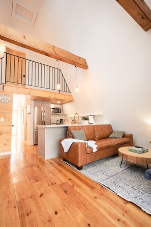 Amplia sala de estar con piso de madera, un sofá de cuero marrón, una mesa de centro redonda y una cocina con electrodomésticos modernos.