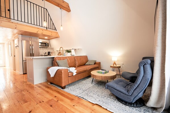 A modern living room with a brown leather sofa, a blue armchair, and a round wooden coffee table. A kitchen with stainless steel appliances is visible in the background.