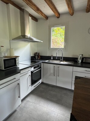 A modern kitchen with white cabinetry, a stainless steel range hood, a microwave, and a window with a view of greenery. - House in the heart of nature (Gabriac)