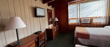 A room with two beds, a desk with a lamp, a dresser, and a television on the wall.