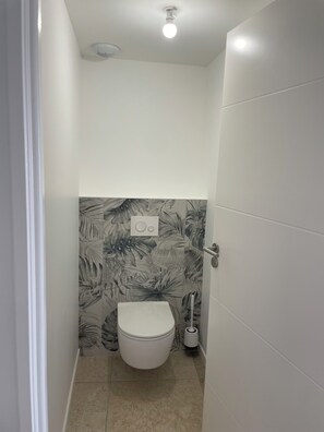 Une petite salle de bain étroite, avec des toilettes, une radio fixée au mur et un luminaire au plafond.