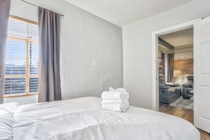 A hotel room with a bed, a stack of towels, a window with blinds, and a door leading to a seating area.