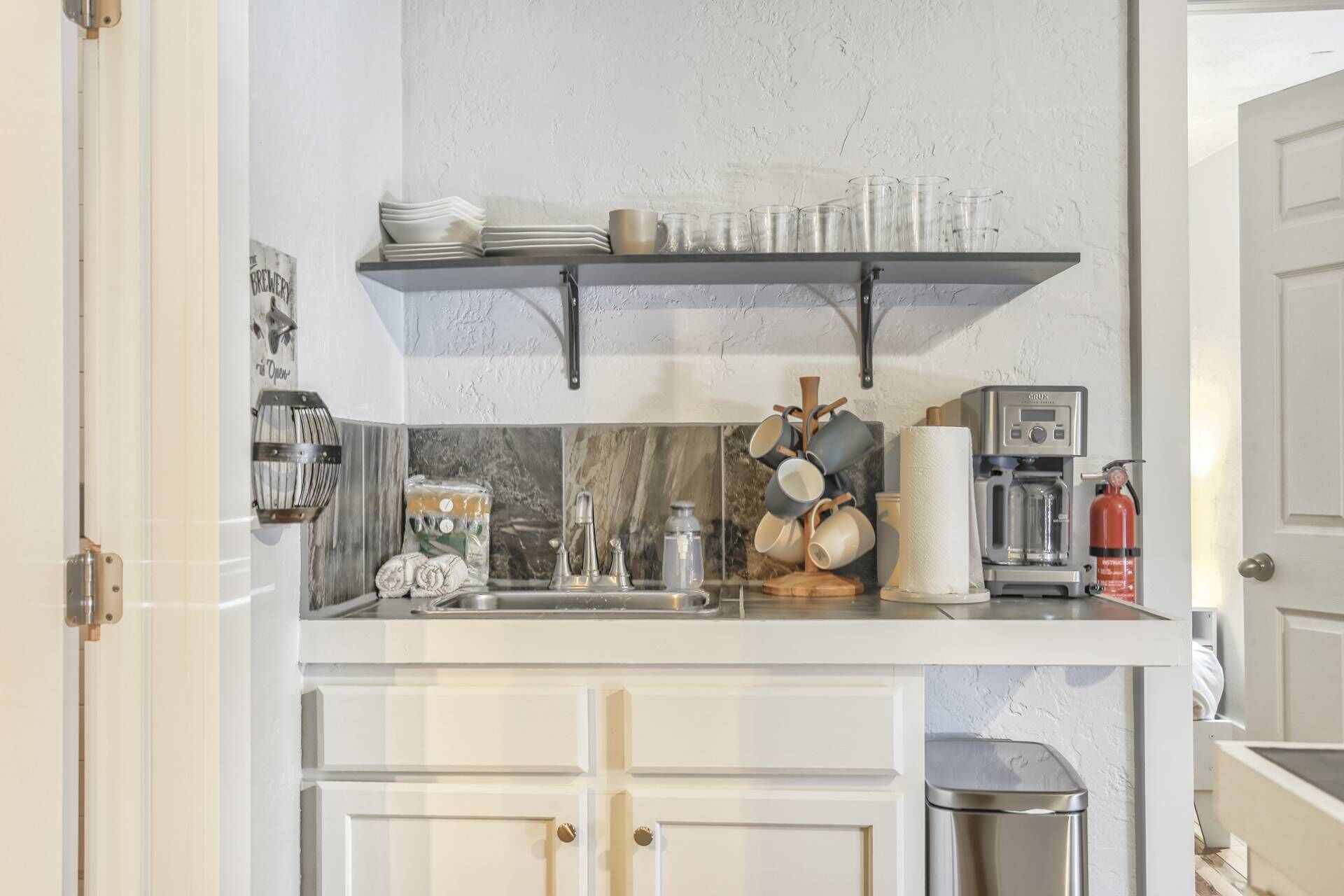 Une cuisine équipée d’une machine à café, d’un extincteur et d’une étagère contenant des assiettes et des verres.