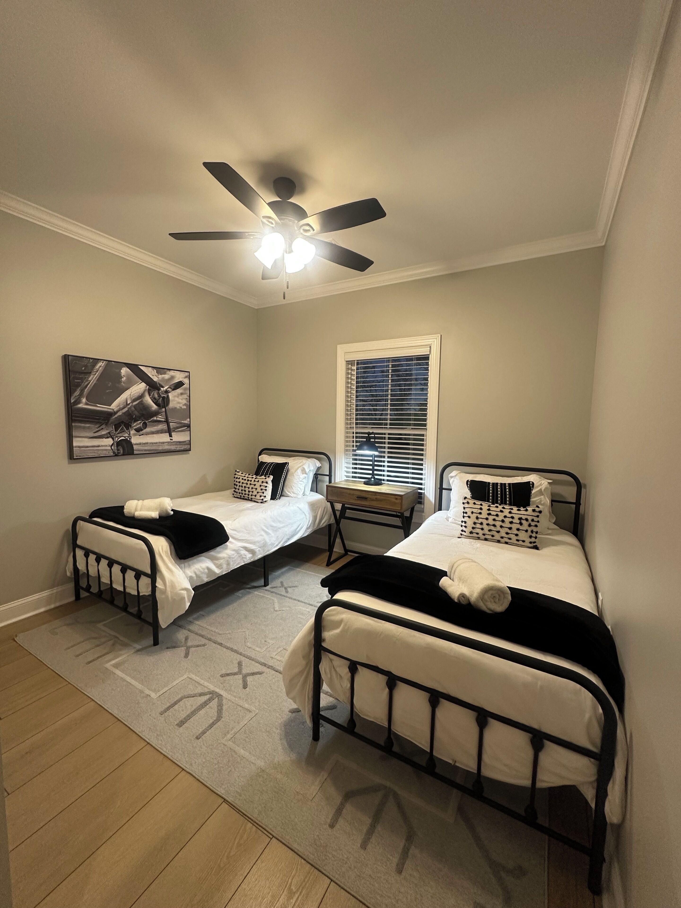 Una camera da letto con due letti, un ventilatore a soffitto, una finestra con persiane e un quadro appeso al muro.