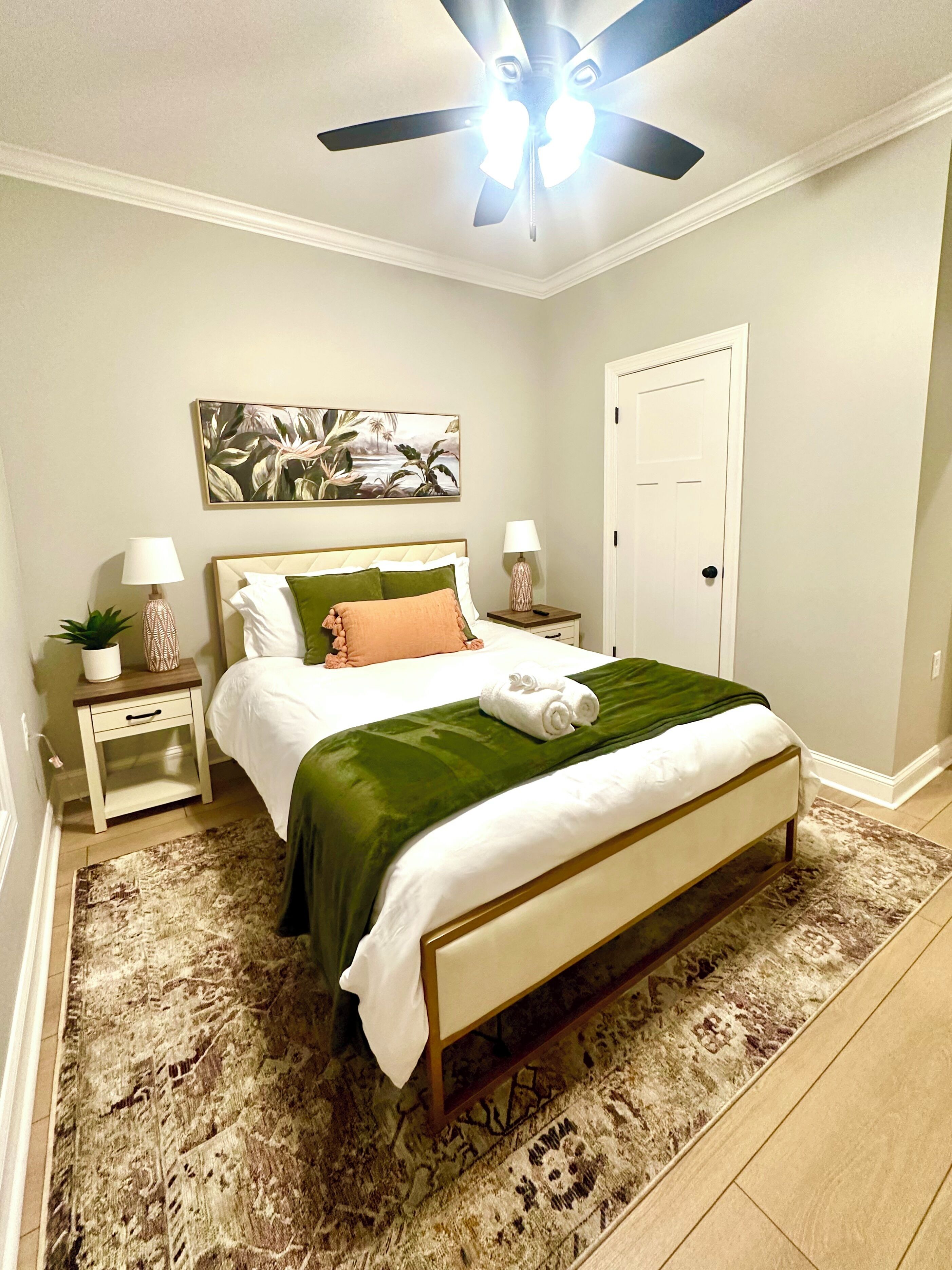 A bedroom with a bed, nightstands, a ceiling fan, a painting, and a door.