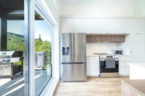 A modern kitchen with stainless steel appliances, a view of greenery outside, and a sliding glass door. - Nature & Mountain | Altisud (Saint-Philémon)