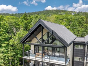 Ein modernes Haus mit einem großen Glasbalkon, umgeben von üppiger Vegetation und einem bewaldeten Hang.