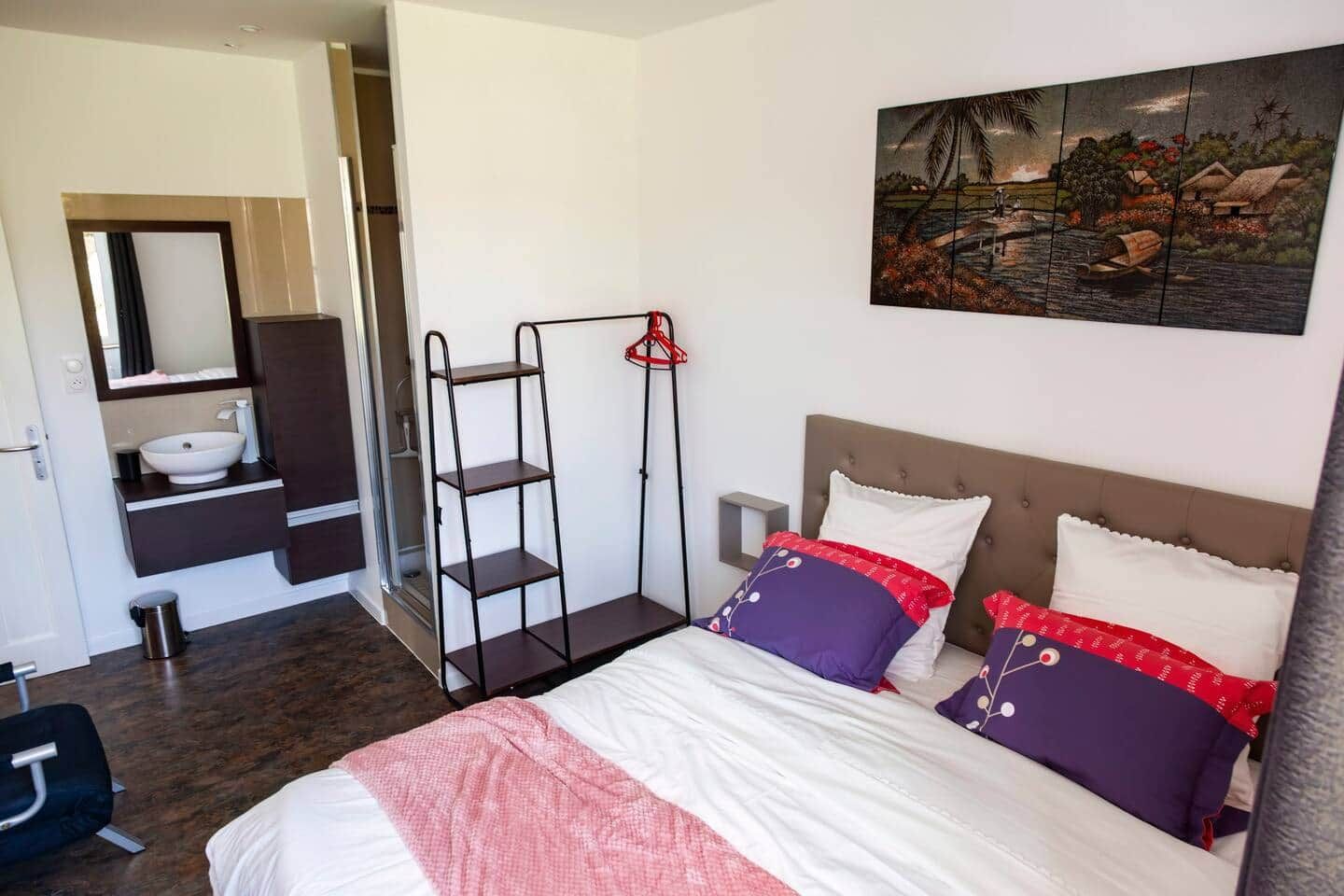A bedroom with a bed, a nightstand, a shelf unit, a painting, and a bathroom with a sink and mirror.