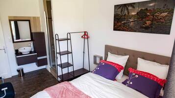 A bedroom with a bed, a nightstand, a shelf unit, a painting, and a bathroom with a sink and mirror.