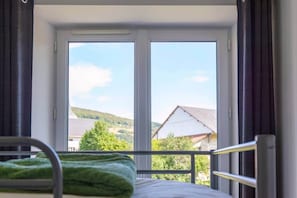 A room with a bed, a window with curtains, and a view of a house and hills.
