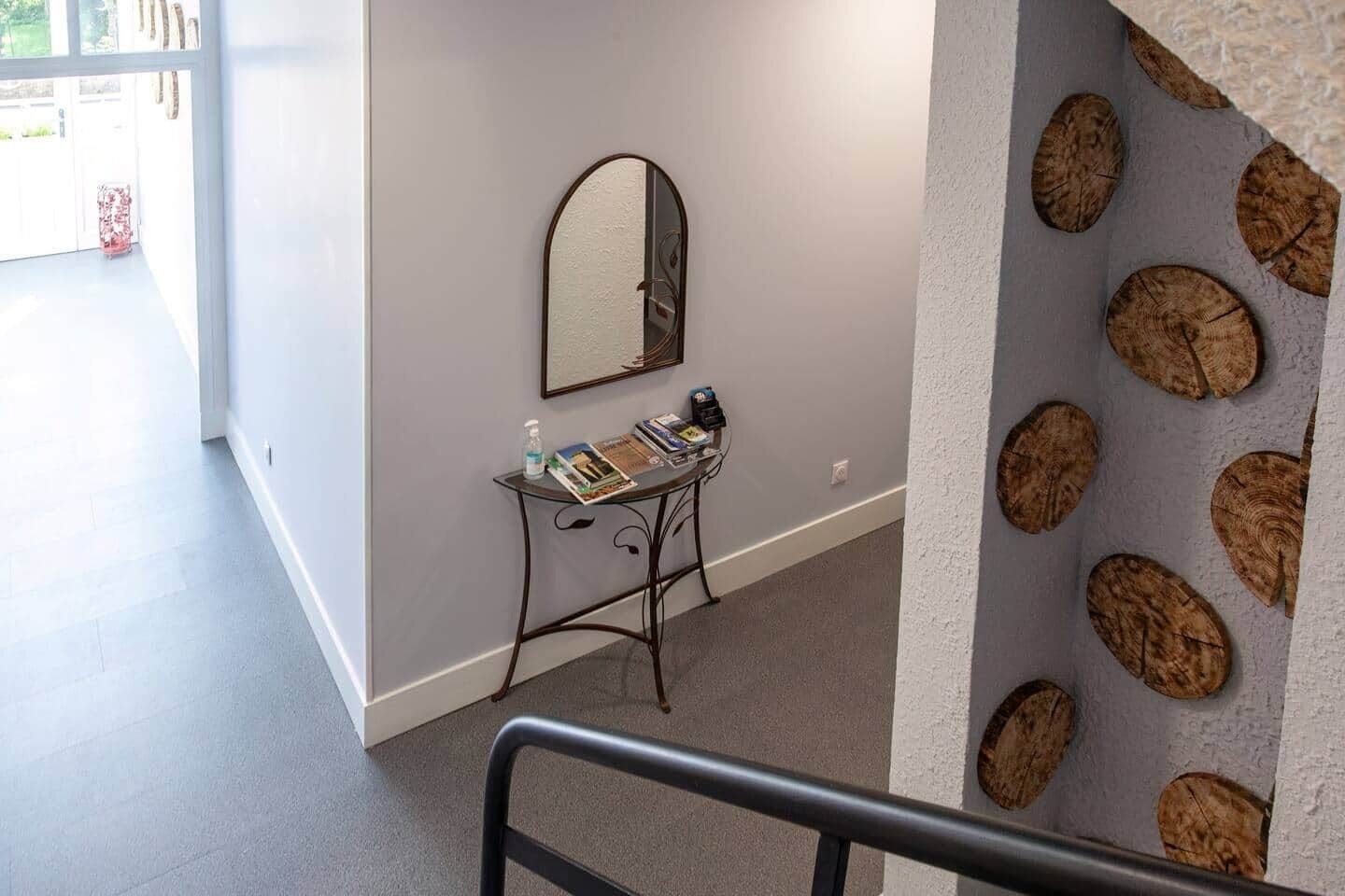 A hallway with a small table holding magazines and a bottle, a mirror on the wall, and a textured wall with circular patterns.