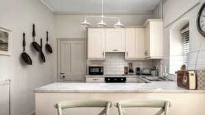 A kitchen with white cabinets, a microwave, a toaster, and a sink. There is a bowl of fruit on the table. - Raphael Rooms (Peterborough)