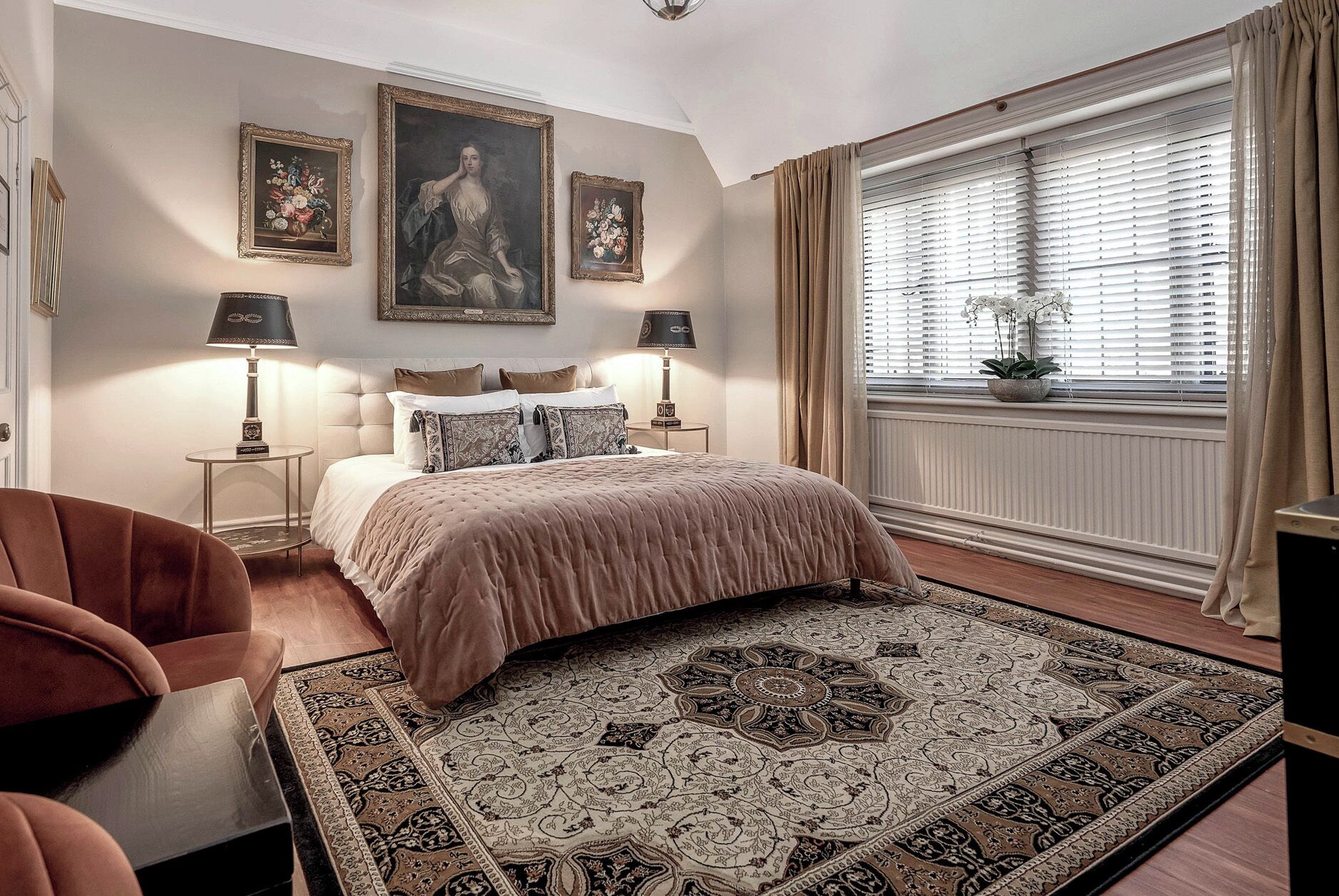 A bedroom with a large bed, two bedside tables with lamps, a chair, a window with curtains, and a wall with framed artwork.