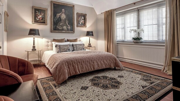 A bedroom with a large bed, two bedside tables with lamps, a chair, a window with curtains, and a wall with framed artwork. - Raphael Rooms (Peterborough)