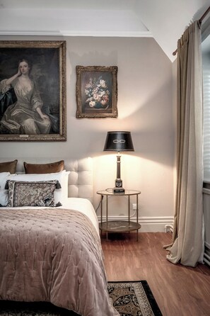 A bedroom with a large bed, a nightstand with a lamp, a framed painting, and a floral picture on the wall. - Raphael Rooms (Peterborough)