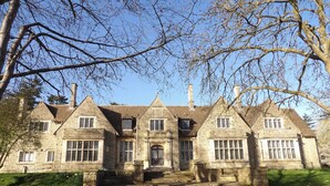 A stone building with multiple windows and a central entrance, surrounded by a grassy area and trees. - Raphael Rooms (Peterborough)