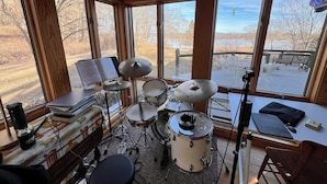A room with a drum set, a keyboard, and a music stand with sheet music. There are large windows with a view of a deck and trees.